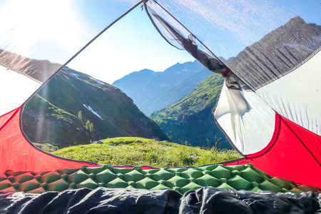 A view through an unfolded tent on a meadow in high Alps. Slopes of the mountains partially covered...