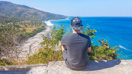 A young man siting on an edge of a stony steep and looking down on a idyllic white sand beach on Lombok, Indonesia. The beach line is overgrown with trees. Calmness and solitude while travelling.の写真素材