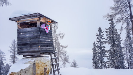 Girl in a skiing outfit and helm standing inside of a hunting pulpit, and leaning through the window. The ski slope is wreathed with fog. The visibility is very low. Girl is happy and enjoying herselfの写真素材