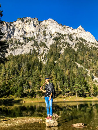 A woman standing on a rock at the shore of Green Lake in an Alpine valley in Austria and enjoying the view. Spring in the valley. High mountain range in the back. Dense forest around the lake.の写真素材