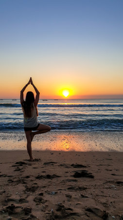A girl practising yoga during the sunset on Seminyak beach on Bali, Indonesia. The sun sets directly into the water. Calm sea washes gently the shore. The sunbeams reflecting on the sea surface.の写真素材