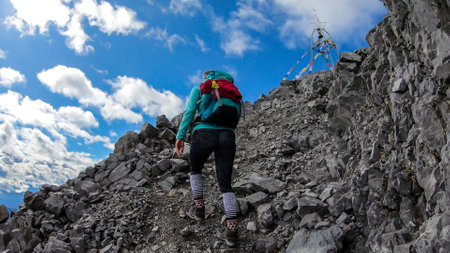 A woman reaching the top of Monte Coglians, Hohe Warte on the Austrian-Italian Alpine border. Metal construction with a bell, wrapped with prayer flags. She is not giving up, visible goal. Overcastの写真素材