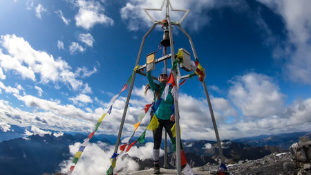 A woman standing under a metal construction with a bell, wrapped with prayer flags at the top of...