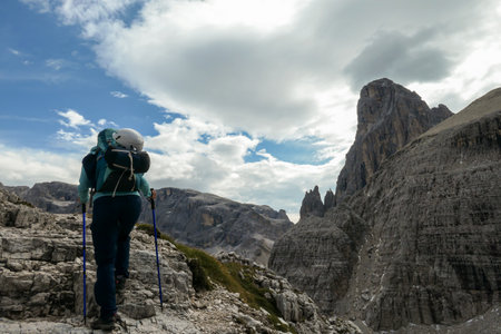 Woman with big backpack and sticks hiking in high Italian Dolomites. She goes up a steep slope. There are many sharp peaks in front of her. She is going up. There are a few trees around. Sunny day.の写真素材