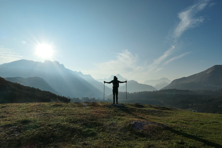 A man with hiking sticks spreading his arms wide open on an early morning in Italian Dolomites. The valley below is shrouded in morning haze. In the back there are high mountain chains. Golden hourの写真素材
