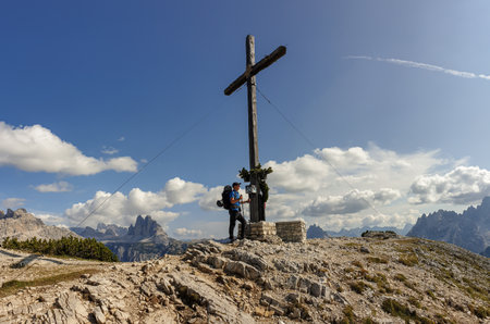 A man with a big hiking backpack standing under a massive wooden cross at the top of  Strudelkopf in Italian Dolomites. Drei Zinnen in the back. The man enjoys the view. High Alpine landscape. Freedomの写真素材