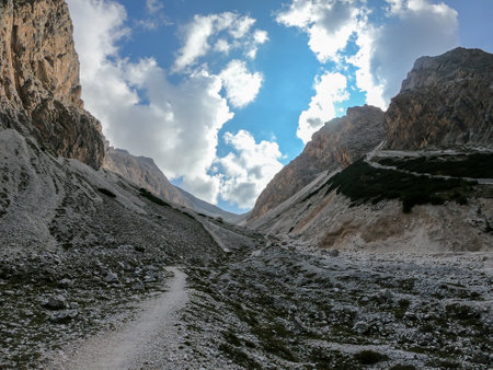 A panoramic view on a stony valley in Italian Dolomites. There are high and sharp mountains around. A narrow pathway leading through the stones. Remote and raw landscape. Sunny day. A bit of overcastの写真素材