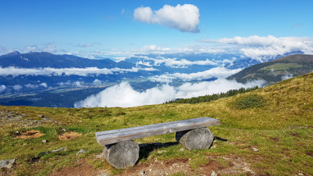 A wooden bench with a panoramic view on the Alpine valley from the top of Granattor in Austrian Alps. The valley is shrouded in fog. The slopes are lush green. Many mountain poking above the cloudsの写真素材