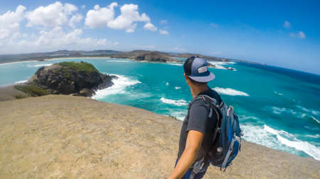 A young man in a full cap walking on a cliff of Tanjung Aan Beach on Lombok, Indonesia and taking a selfie. Sea water is calm, shimmering with many shades of blue. Solo traveller in an unspoiled areaの写真素材