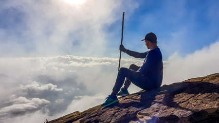 A young man sitting on the side of volcano Inerie in Bajawa, Flores, Indonesia. He is surrounded by clouds, as if he was walking on them. Clear blue sky. Natural phenomenon. He hold a walking stickの写真素材