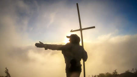 A young woman standing on the top of volcano Inerie in Bajawa, Flores, Indonesia, next to a cross....