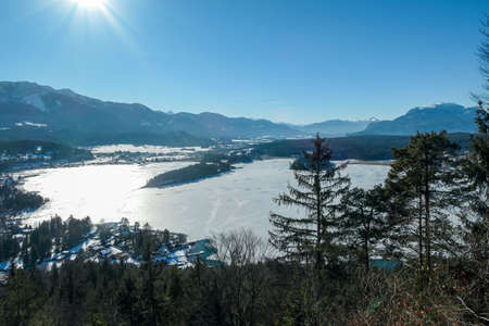 A panoramic view on the Faaker lake in Austrian Alps in winter. The lake is surrounded by high mountains and tall trees at its shore. The surface of the lake is frozen. Clear, blue sky. Frosty winterの写真素材