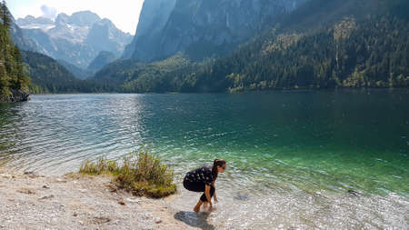 A woman squatting by the shore of Gosau lake and sprinkling the water with her arms. The lake is surrounded by high mountains, with Dachstein glacier in the back. The woman is having fun. Happy momentの写真素材
