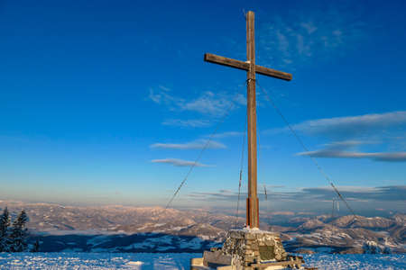 A huge wooden cross on top of Schoeckl, Austrian Alps during the morning golden hour. The peak is covered with powder snow. The mountain chains in the back are turned golden. Soft sunrise colors.の写真素材