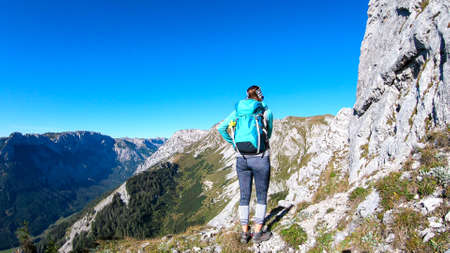 A woman with a hiking backpack climbing to the of a big boulder on the way to Hohe Weichsel in Austria, with a panoramic view on a vast valley.  Narrow pathway. She is enjoying the view. Discoveringの写真素材