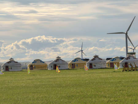 Yellow and white traditional yurts located on a pasture in Xilinhot in Inner Mongolia. Endless grassland. Blue sky with a few thick clouds. Wind turbines in the back. Clean energy. Nomadic way of lifeの写真素材