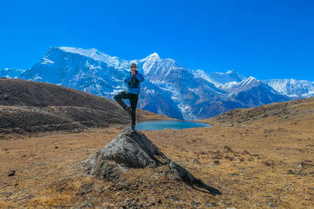 A young man wearing a blue jacket, taking a tree pose at a huge rock, tries to meditate next to Ice Lake, Annapurna Circuit Trek, Himalayas, Nepal. Yogi's remedy. Solitude and awareness.の写真素材