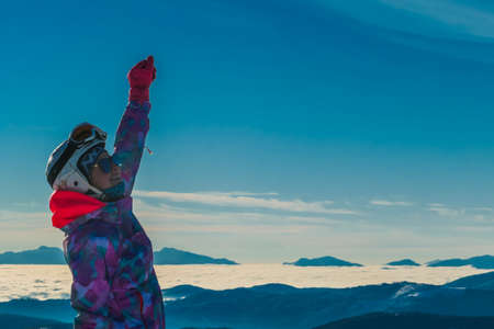 Snowboarder Girl with a hand raised up, Moelltaler gletscher, Austria. Valley covered with clouds. She wears colorful jacket, sunglasses and helmet. Smiley face shows content and happiness.の写真素材