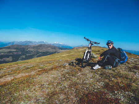 A young boy on a mountain bike tour in Millstatt, Austria. Boy wears a big backpack and a helmet. Mountain bike stands next to him. Beautiful mountain view in the back. Professional sport gear.の写真素材