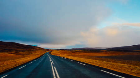 An endless road through the lowlands. On the side some hills. Dark and heavy tones. Both sides of the road are barren. Sky is clouded, but still blue parts visible. Empty road, not a single car movingの写真素材