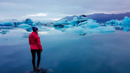 A young woman standing on the rock in the middle of the glacier lagoon. Huge and massive ice bergs...