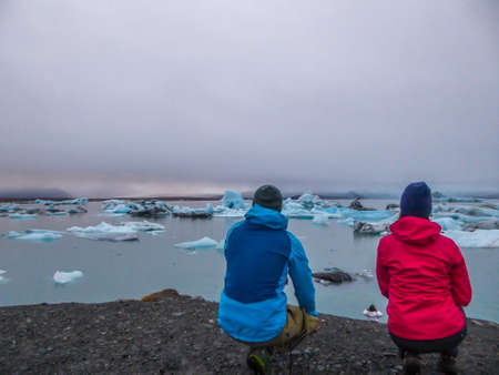 A couple squatting at the edge of a cliff, looking at the glacier lagoon. Girl wearing pink jacket,...
