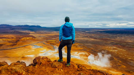 Young man wearing a blue jacket stands on top of a mud mountain, overlooking a geothermal spot noted for its bubbling pools of mud & steaming fumaroles emitting sulfuric gas. Power of planet Earth.の写真素材
