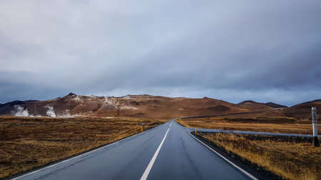 An endless road through the lowlands. In the back tall mountains. Dark and heavy tones. Both sides of the road are barren, overgrown with grass. Sky is clouded. Empty road, not a car passing by.の写真素材