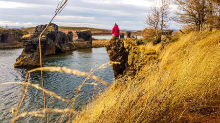 A young woman wearing ink jacket sits at the cliff, overlooking some other islands. Dry grass on the shores of the cliff. Romantic capture of a tender moment of coexistence of human and nature.の写真素材