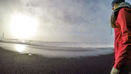 A girl wearing pink jacket walks around the black sand beach. In the back the sun slowly starts setting. Waves push the foamed waves to the shore. White foam contrasted with black sand. Solitudeの写真素材