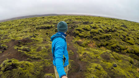 A young man wearing blue jacket and a beanie walks around the lava fields, overgrown with moss,...
