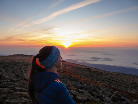 A woman with a headband sitting on top of Babia Gora, Poland, and enjoying the panoramic view on sun rising above the horizon. There are thick clouds below. The sky is pink and orange. Calmnessの写真素材