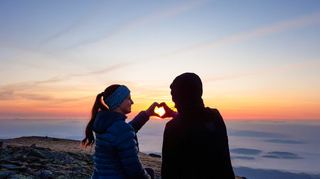 A couple sitting on top of Babia Gora, Poland, and enjoying the panoramic view on sun rising above the horizon. They form a heart with their hands. Thick clouds below. The sky is pink and orange. Loveの写真素材