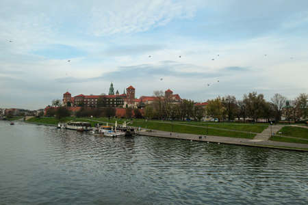 A panoramic view on the iconic Wawel Castle in Cracow, Poland and the Vistula river flowing under the castle. A few birds flying above the river. A few boats docked on the rivers bend. City tour.の写真素材