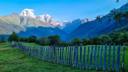 A wooden fence in front of a lush pasture with a view on the first sunbeams reaching the peaks of Ushba in Caucasus, Georgia. Cloudless sky above the high and snow-capped mountains. Daybreak.の写真素材