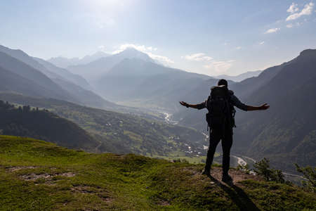 A man with big hiking backpack spreading his arms wide open, while standing on the mountain peak,...