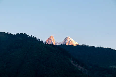 First sunrise reaching the peaks of Ushba in Caucasus, Georgia. Cloudless sky above the high and snow-capped mountains. The hills below are shrouded with shadows. Daybreak. High and desolate mountainsの写真素材