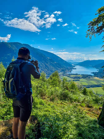 A man taking pictures at a viewing point near the top of Oswaldiberg with the view on the Lake Ossiach in Austrian Alps, Carinthia. The photographer enjoys a panoramic view on the Ossiacher Tauern.の写真素材