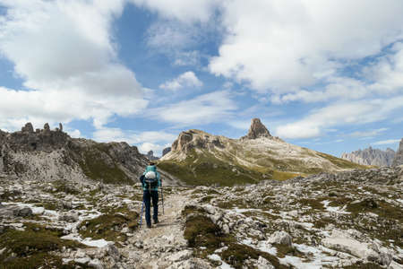 A woman with backpack and sticks hiking on a narrow path in Italian Dolomites. There are sharp and steep mountains. At the bottom of a small valley there is a small, navy blue lake. Raw landscape.の写真素材