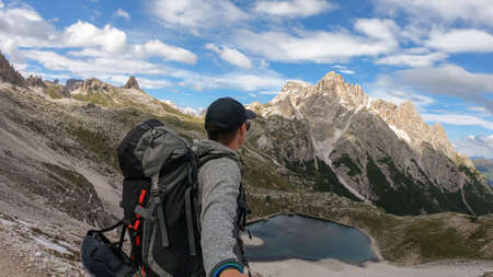 A man with hiking backpack and sticks taking a selfie in Italian Dolomites. There are sharp and steep mountains. At the bottom of a small valley there is a small, navy blue lake. Raw landscape. Remedyの写真素材