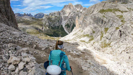 A woman with backpack and sticks hiking on a narrow path in Italian Dolomites. There are sharp and steep mountains around her. Lots of lose stones. Raw and desolated landscape. Following the pathwayの写真素材