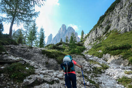A woman with big hiking backpack hiking in high Italian Dolomites. There are many sharp peaks behind. She walks on a very narrow pathway. There are a few trees around. Sunny day. Outdoor exercisingの写真素材