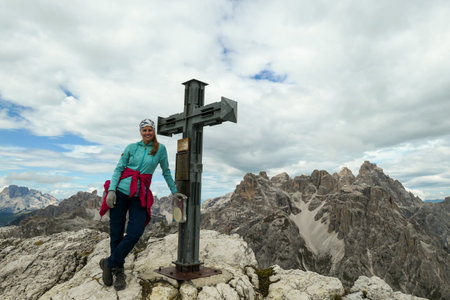A woman standing next to a wooden cross on high and desolated mountain peak in Italian Dolomites. In the back there are endless mountain chains. Few clouds above the peaks. Sharp and steep slopes.の写真素材