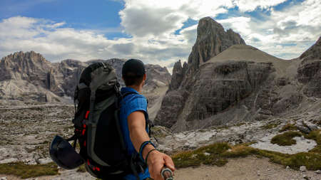 A man taking a selfie while hiking in Italian Dolomites. He has a big backpack. There are endless mountain chains in front of him. The high peaks are very stony and sharp. Discovering remote places.の写真素材