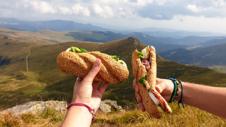 Lunch break in Austrian Alps. Two hands holding sandwiches with the view on high Alps. There are endless mountain chains in the back. Eating in the fresh air. A bit of overcastの写真素材