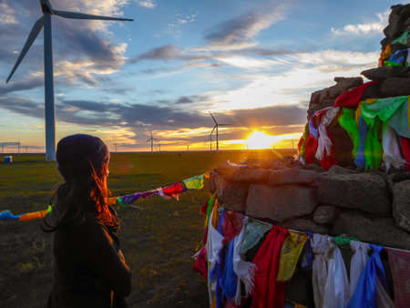 A woman in beanie walking around a Heaps of stones (Aobao) build on a vast pasture in Xilinhot in Inner Mongolia. The Heaps has colorful prayer flags attached to it. The sun is setting. Wind turbinesの写真素材