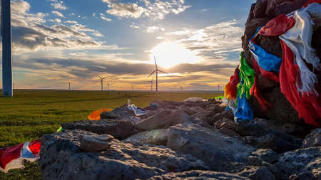 Heap of stones (Aobao) build on a vast pasture in Xilinhot in Inner Mongolia. The heap has colorful prayer flags attached to it. The sun is setting behind the horizon. Golden hour. Wind turbinesの写真素材