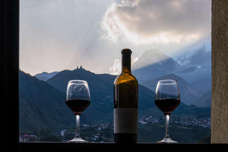 Georgian red wine with a panoramic evening on Mount Kazbegi in the Greater Caucasus Mountains in Georgia. Clouds are covering the snow-capped summit. Sunset, sunrise. Romantic date, two wine glasses.の写真素材