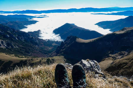 A hiker having a break on the summit of Eisenerzer Reichenstein in Styria, Austria, Europe. The Ennstal valley is covered in clouds and fog. Hiking trail, Wanderlust. Sunny day. Hiking shoe. Freedomの写真素材