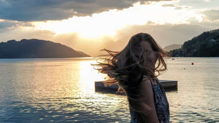 A woman in a blue dress flips her long, brown hair by the shore of Millstaettersee lake. The sun is...